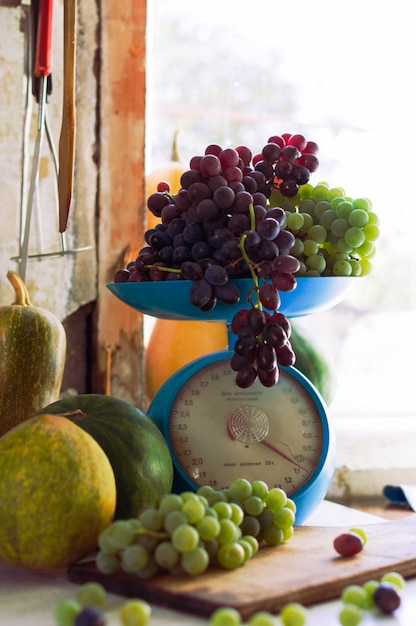 Herbststillleben mit Kürbissen, Melonen, Wassermelonen, Trauben auf einer Waage und in einer Metallschale auf einem weißen Holztisch. Herbsterntekonzept.