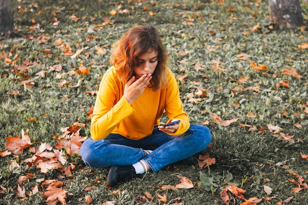 Herbststimmung. Junges Mädchen im gelben Pullover mit Smartphone, das auf Herbstgras mit trockenen Blättern sitzt