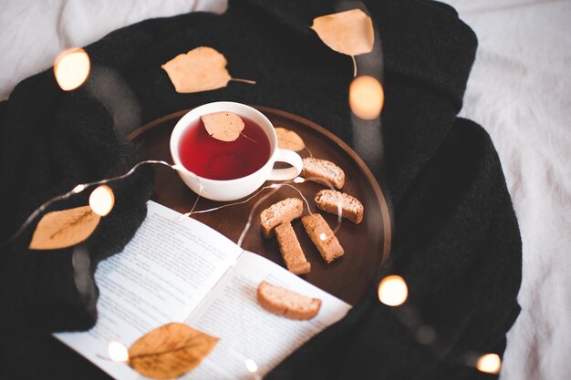 Herbsttasse roter Tee mit gefallenen Blättern und offenem Buch auf Strickpullover im Bett. Guten Morgen. Herbstsaison. Frühstück.