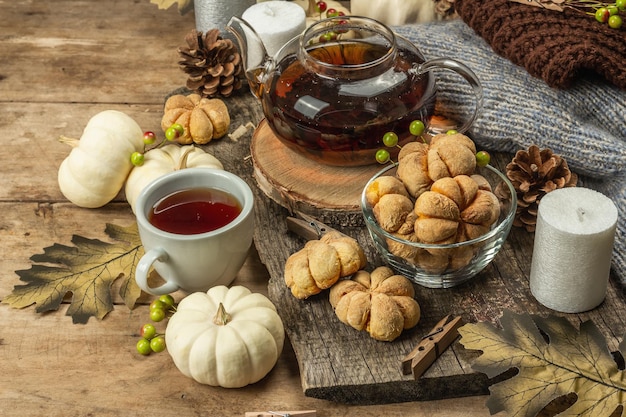 Herbsttee-Konzept. Kekse mit Kürbispüree, schwarzer Tee in einer Glasteekanne, Herbstdekor. Kranz, Kerzen, ein kuscheliger Pullover. Alter hölzerner Hintergrund, Nahaufnahme