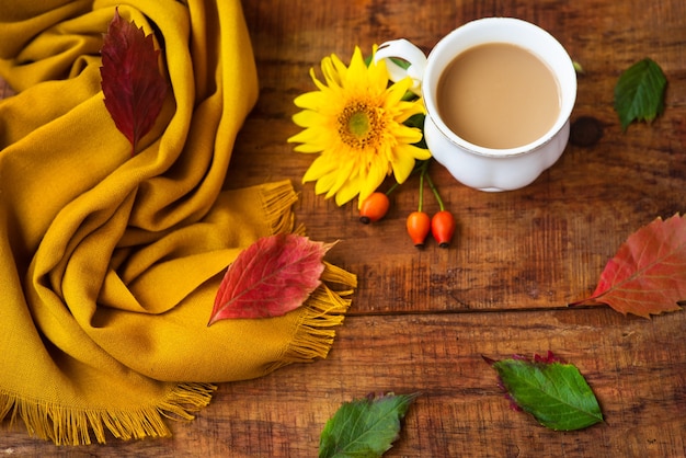 Herbsttee-Tassenzusammensetzung, gelber Schal, Rosenbeeren und Sonnenblumenblume auf einem hölzernen Hintergrund. Herbst Hintergrund. Warme, gemütliche Atmosphäre des Herbstes. Flache Lage, Layout