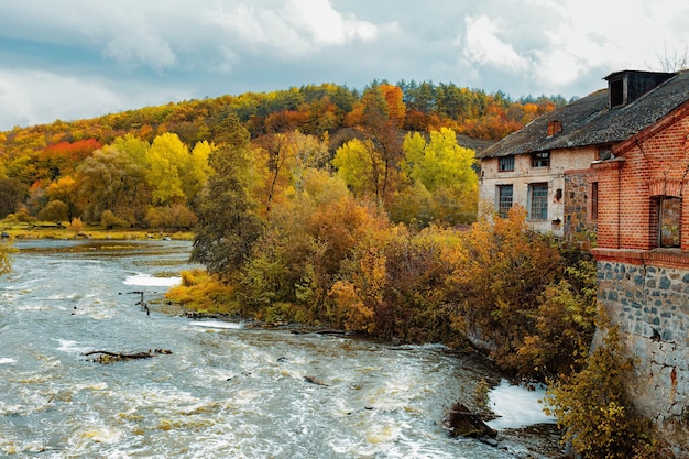 Herbstwald am Flussufer, verlassenes Steingebäude, blauer Himmel und weiße Wolken