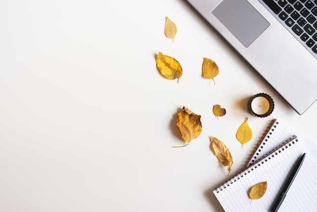 Herbstwohnung mit Laptopkerzennotizbüchern und getrockneten gelben Blättern auf weißem Schreibtisch