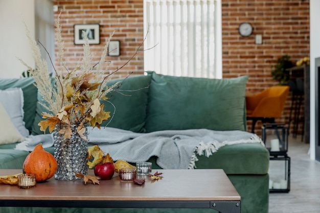 Herbstzeit dekoriertes zuhause. gelber blumenstrauß in glasvase auf stilvollem holztisch, kürbis, rote äpfel, blätter, kerzen, decke. grünes sofa, backsteinmauer und kamin im hintergrund