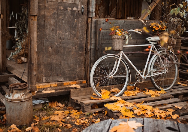 Herbstzeit. Vintage-Fahrrad mit bunten Blumen im Korb, der sich während der Herbstsaison an die Holzwand des alten stimmungsvollen Landhauses lehnt, Schnurbeutel mit Herbsternte an der Tür hängend