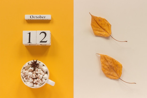 Herbstzusammensetzung. Holzkalender 12. Oktober, Tasse Kakao mit Marshmallows und gelbem Herbstlaub auf gelb-beigem Hintergrund. Ansicht von oben Flach Mockup-Konzept Hallo September.