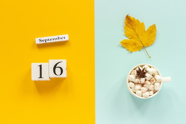 Herbstzusammensetzung. Holzkalender 16. September, Tasse Kakao mit Marshmallows und gelbem Herbstlaub auf gelbblauem Hintergrund. Ansicht von oben Flach Mockup-Konzept Hallo September.