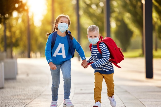 Hermana y hermano encantados con ropa informal y máscaras protectoras con mochilas tomándose de la mano y corriendo juntos en el parque listos para los estudios escolares