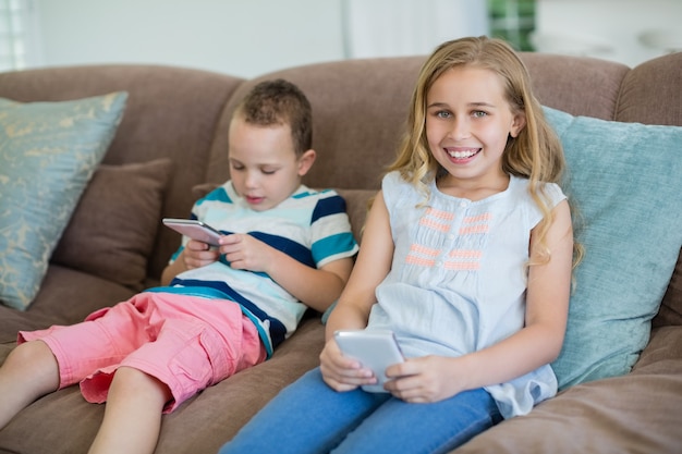 Hermana y hermano sonriente sentado en el sofá mediante teléfono móvil en el salón