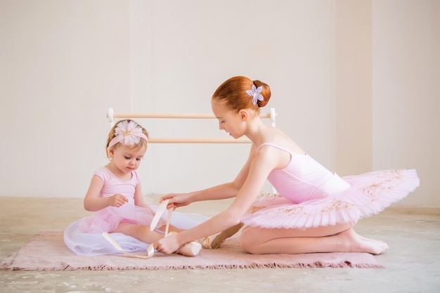 La hermana mayor, una bailarina con un tutú rosa, le pone zapatos de punta al bebé mientras se sienta en la barra