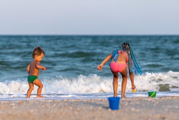 Una hermana mayor linda y flaca juega con su hermano pequeño mientras se sienta en el agua poco profunda del mar cerca de la orilla, en un cálido día soleado en las vacaciones de verano