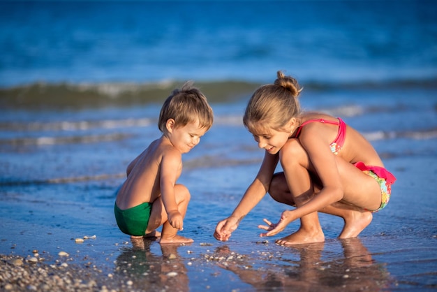 Una hermana mayor linda y flaca juega con su hermano pequeño mientras se sienta en el agua poco profunda del mar cerca de la orilla, en un cálido día soleado en las vacaciones de verano