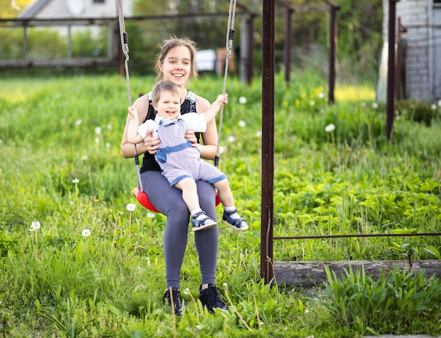 La hermana mayor lleva ropa oscura de primavera y su hermano menor es un niño pequeño con un mono brillante. Ella lo monta en un columpio rojo y disfrutan de la primavera en un jardín floreciente en un clima cálido de primavera.