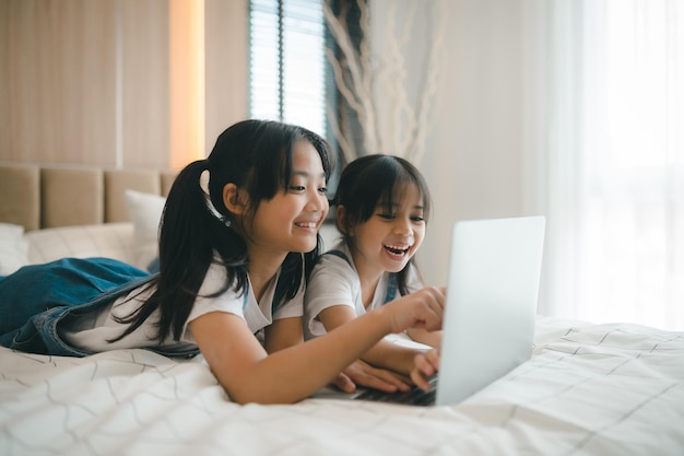 Hermana niña asiática en la cama y aprendiendo en línea en una computadora portátil Internet Lección de clase virtual en video durante el autoaislamiento en el hogar Educación remota remota en video Estudio escolar moderno para niños