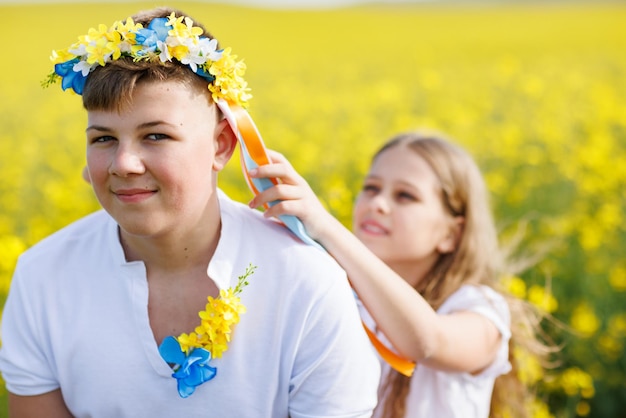 Hermana trenza cintas en corona ucraniana en la cabeza de su hermano adolescente contra el telón de fondo de los campos y el cielo