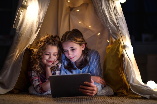 Hermanas adentro en casa, usando tableta en pijama en carpa. Concepto de bloqueo.