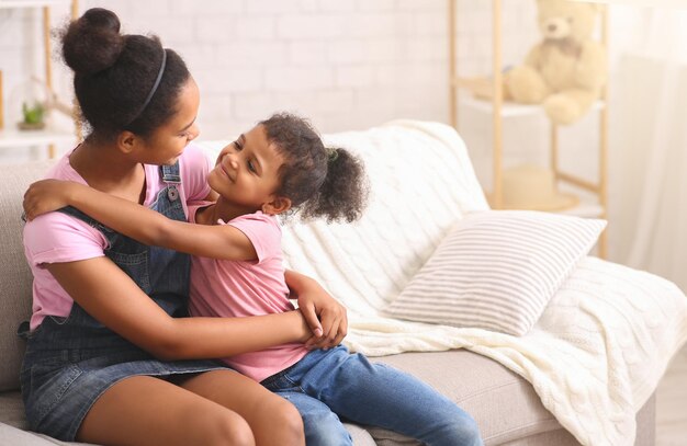 Hermanas africanas felices abrazándose en casa sentadas en el sofá