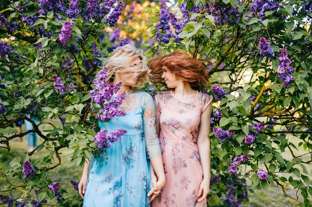 Hermanas gemelas expresivas en hermosos vestidos sacudiendo sus cabezas en el floreciente parque de verano.