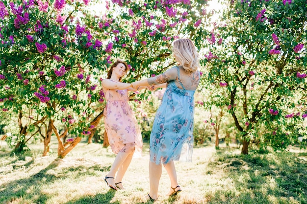 Hermanas gemelas felices lindas en hermosos vestidos girando en el floreciente parque de verano.