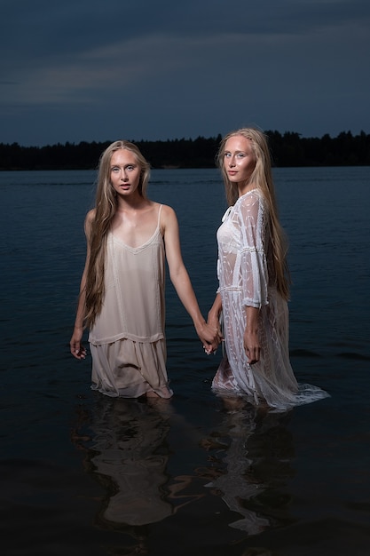 Hermanas gemelas jóvenes con largos cabellos rubios posando en vestidos ligeros en el agua