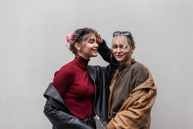 Hermanas hermosas divertidas niñas en ropa de moda con una chaqueta de cuero y un suéter de punto de pie cerca de una pared gris al aire libre