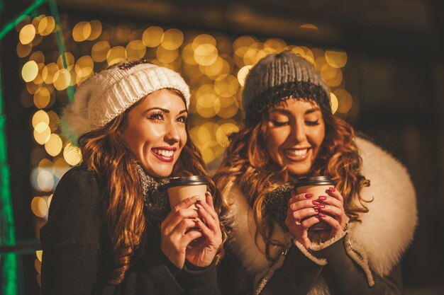 Hermanas jóvenes alegres con tazas de café para divertirse en la noche de invierno en la calle de la ciudad.