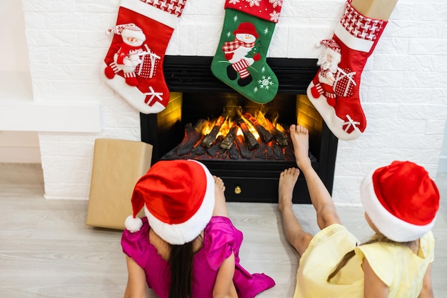 Hermanitas felices junto a una chimenea en una acogedora sala de estar en la víspera de Navidad. Celebrando la Navidad en casa.