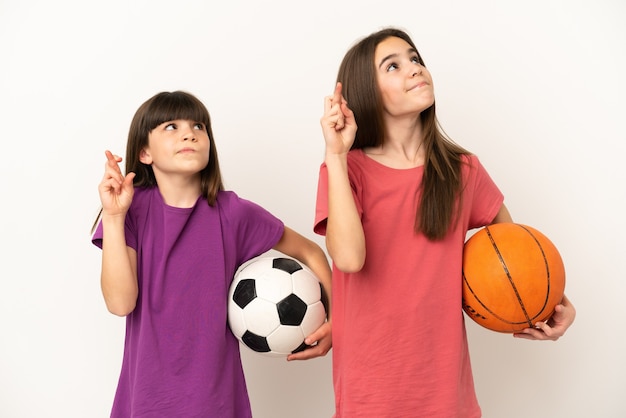Foto hermanitas jugando al fútbol y baloncesto aislado sobre fondo blanco con los dedos cruzando y deseando lo mejor