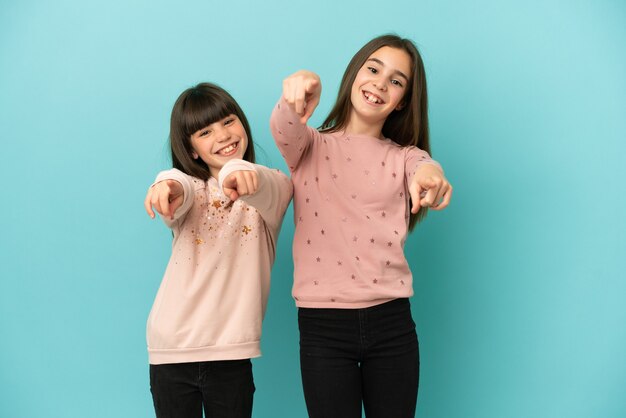 Hermanitas niñas aisladas sobre fondo azul le señala con el dedo mientras sonríe