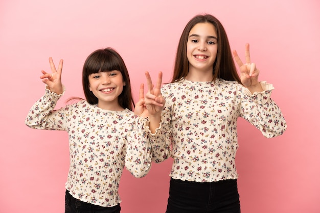 Hermanitas niñas aisladas sobre fondo rosa sonriendo y mostrando el signo de la victoria con ambas manos