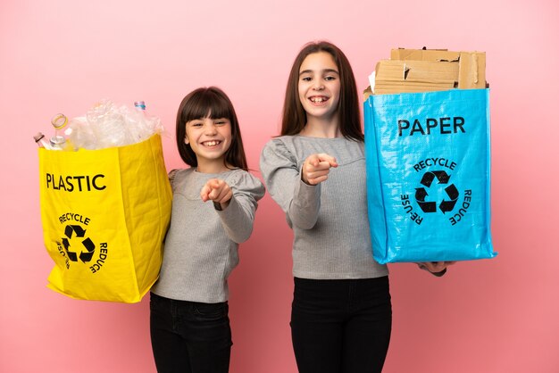 Hermanitas reciclando papel y plástico aislado sobre fondo rosa te señala con el dedo con una expresión de confianza