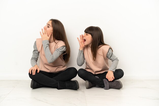 Hermanitas sentadas en el suelo aislado sobre fondo blanco gritando con la boca abierta hacia el lateral