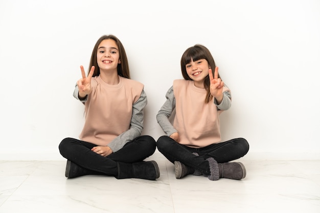 Hermanitas sentadas en el suelo aislado sobre fondo blanco sonriendo y mostrando el signo de la victoria