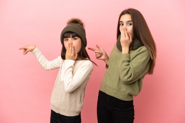 Hermanitas vistiendo una ropa de invierno aislado sobre fondo rosa dedo acusador hacia el lado con una cara de sorpresa