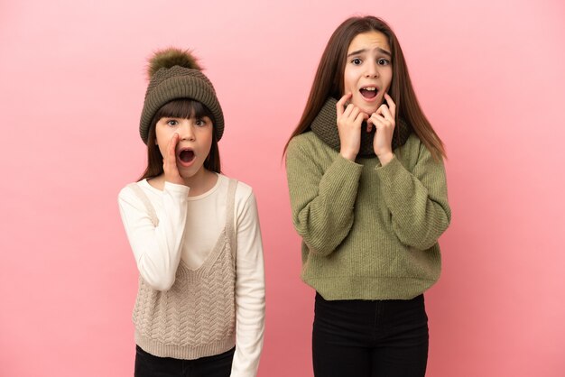 Hermanitas vistiendo una ropa de invierno aislado sobre fondo rosa sorprendido y conmocionado mientras mira a la derecha