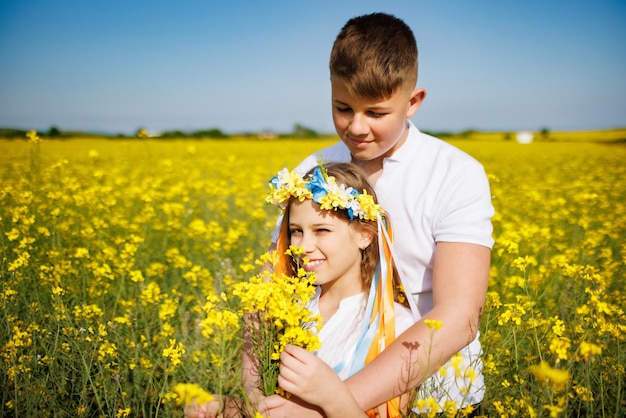 Hermano abraza a hermana con corona ucraniana y ramo de flores en las manos en el campo de colza bajo el cielo