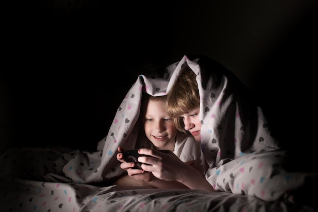 Hermano y hermana se acuestan debajo de una manta por la noche y juegan por teléfono