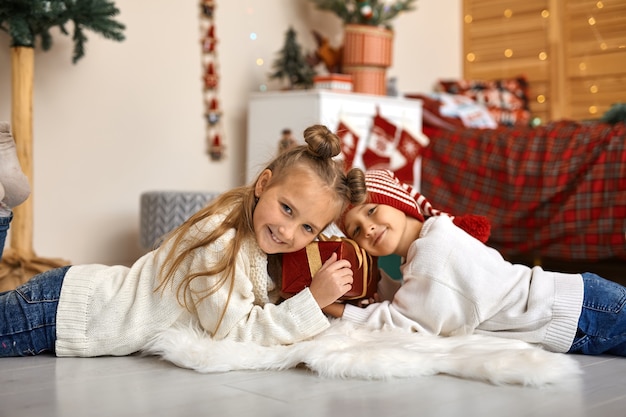 Un hermano y una hermana alegres yacen en el suelo de la habitación de sus hijos decorada para la Navidad y sostienen juntos una hermosa caja de regalo.