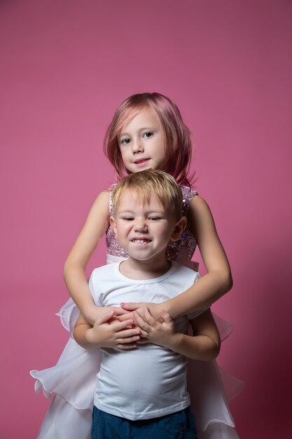 Hermano y hermana caucásicos, abrazándose a la cámara en tiro de estudio de fondo rosa. Lazos familiares, amistad, concepto de infancia feliz.