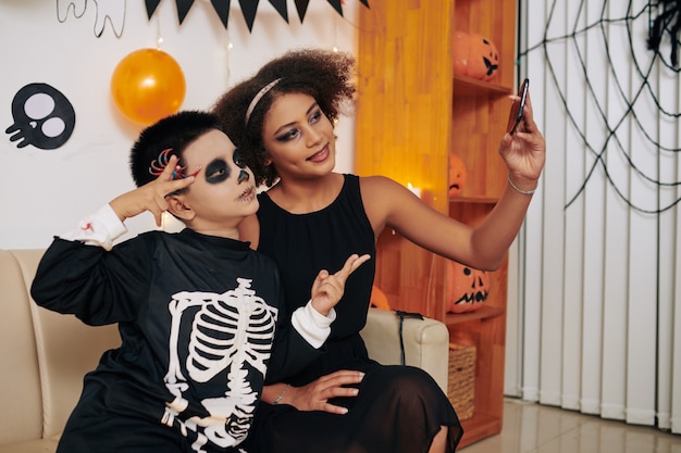 Foto hermano y hermana hablando de fotos en el teléfono inteligente o videollamadas a amigos en la fiesta de halloween en casa