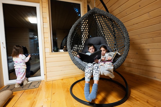 Hermano con hermana leyendo libros en una cómoda silla colgante en una acogedora cabaña de madera pequeña Vida en el campo