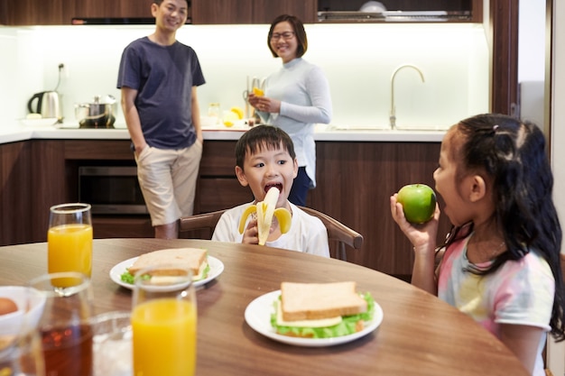 Hermano y hermana mirándose y comiendo frutas para el desayuno en la cocina, sus padres de pie en segundo plano.