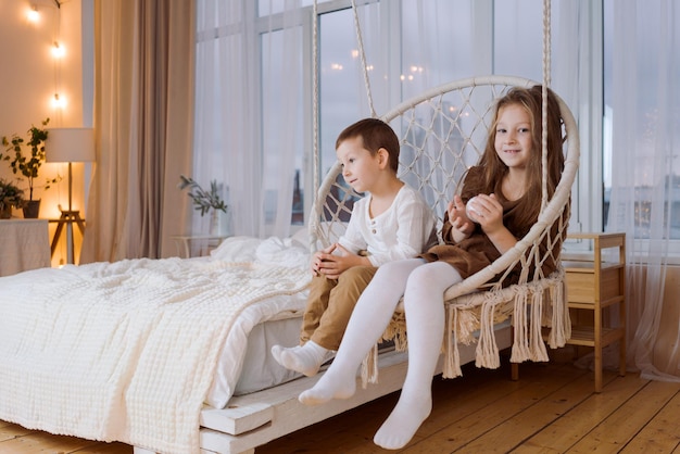 El hermano y la hermana pequeños viajan en una silla colgante de estilo escandinavo en el dormitorio