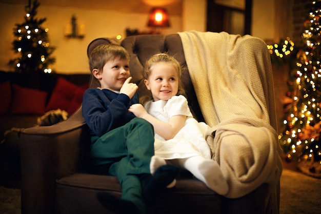 Hermano y hermana posando en un sillón en un acogedor interior navideño con árbol de navidad