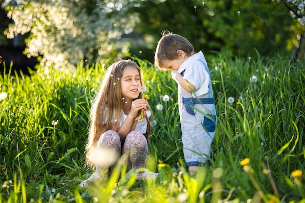 Hermano y hermana en ropa de verano brillante. Divertido y divertido juego con esponjosos dientes de león blancos y amarillos con el telón de fondo de hierba alta y frondosos árboles verdes en el jardín de primavera.