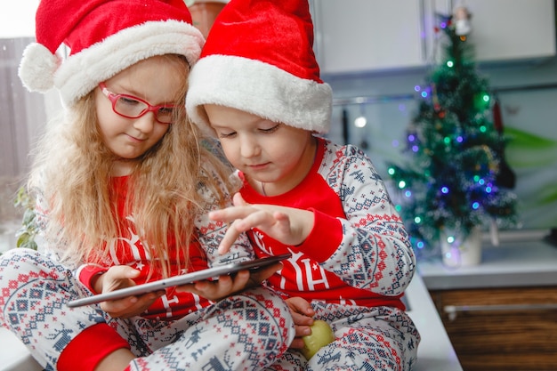 Hermano y hermana se sientan en la cocina con gorros de Papá Noel y miran la libreta. Los niños escriben una carta a Santa Claus