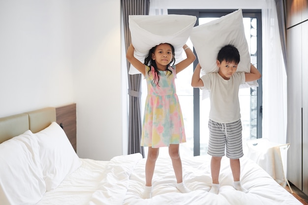 Hermano y hermana vietnamita jugando con almohadas en la cama en el dormitorio de los padres y mirando a la cámara