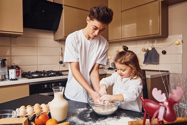 Hermano con su hermana pequeña preparando comida usando harina en la cocina y diviértete.