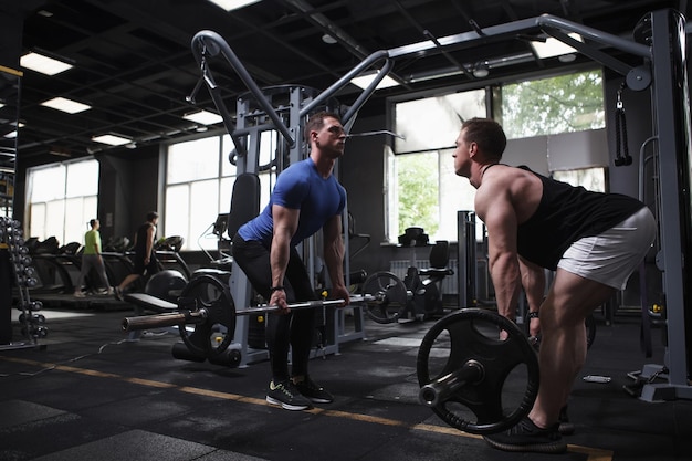 Foto hermanos gemelos culturistas haciendo ejercicio de peso muerto con barra de pie cara a cara en el gimnasio