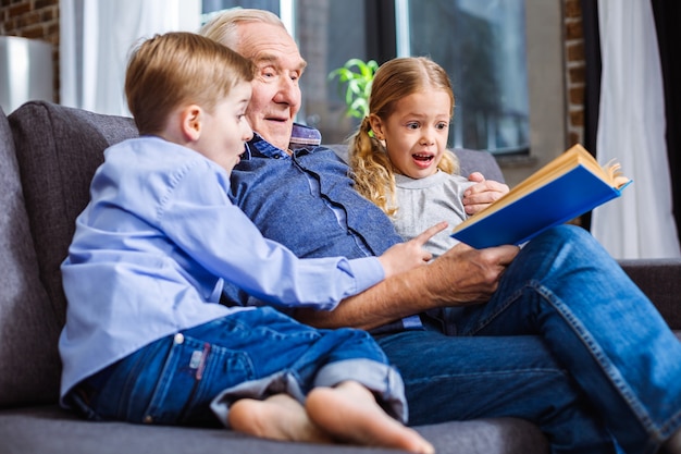 Hermanos pequeños alegres que leen un libro mientras descansan con su abuelo en el sofá